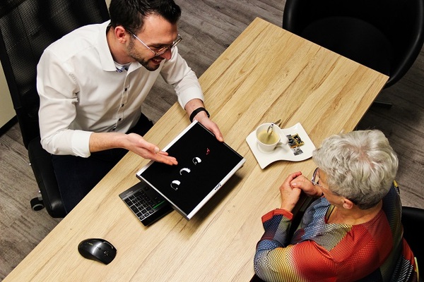 Ein Hörakustiker berät eine Seniorin am Tisch zu verschiedenen modernen Hörgeräten.